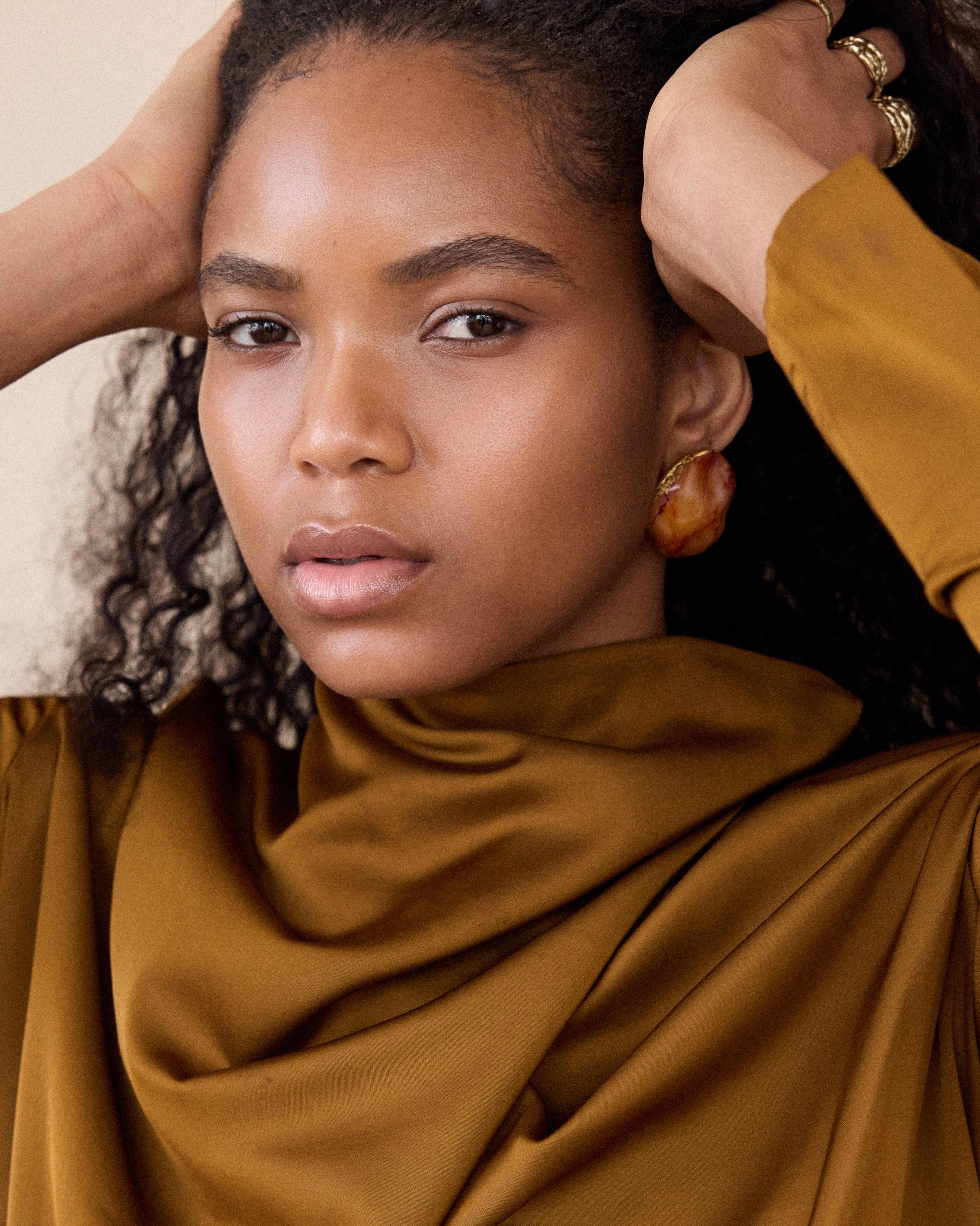 Woman wearing a mustard yellow top with hands on her head against a neutral background